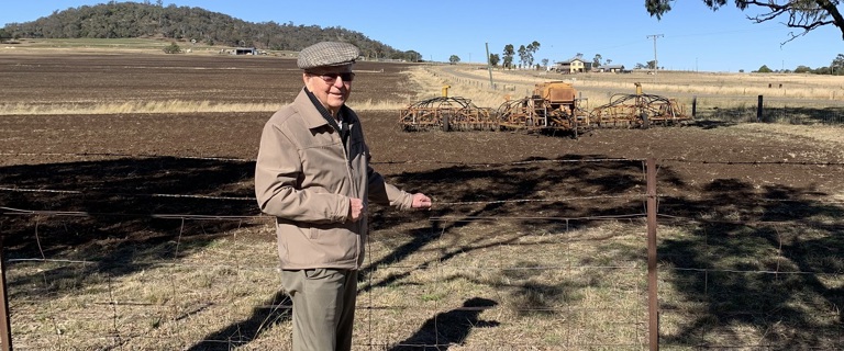 An elderly gentleman stands in front of a paddock where a seeder can be seen in the background