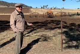 An elderly gentleman stands in front of a paddock, a seeder machine can be seen in the background